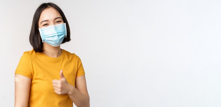 Covid-19, vaccination and healthcare concept. Portrait of cute asian girl in medical mask, has band aid on shoulder after coronavirus vaccine, standing over white background.