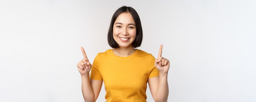 Portrait of beautiful japanese girl smiling, pointing fingers up, showing advertisement, standing in yellow tshirt against white background.