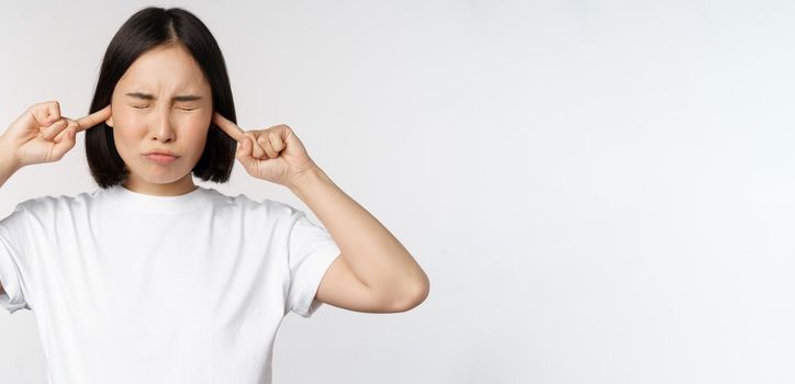 Portrait of asian woman shut ears and feeling discomfort from loud noise, annoying sound, standing over white background.