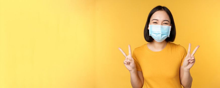 Positive asian woman smiling, wearing medical face mask from covid-19 during pandemic, showing peace v-sign gesture, yellow background.