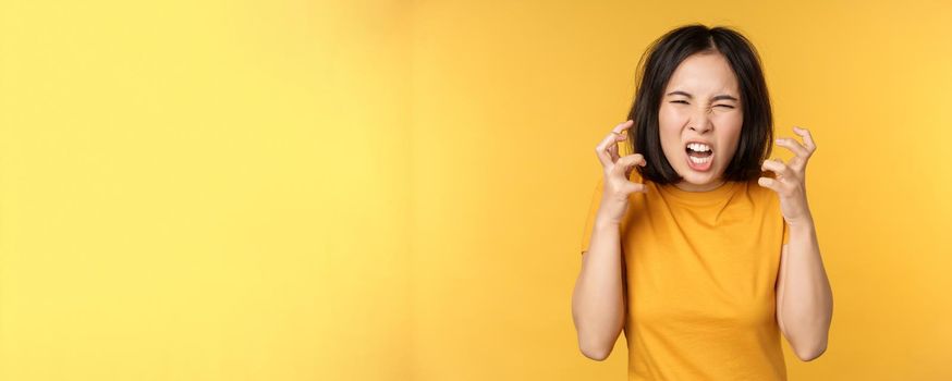Image of angry asian woman, shouting and cursing, looking outraged, furious face expression, standing over yellow background.