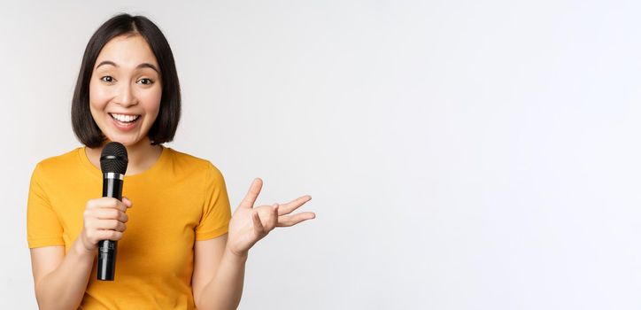 Image of young asian woman talking in microphone, perfom with mic, giving speech, standing in yellow tshirt against white background.