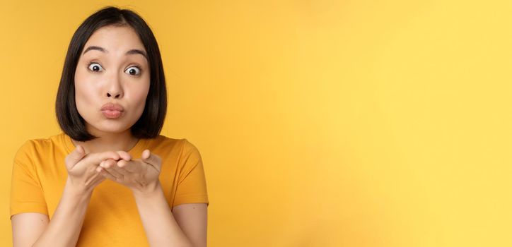 Cute asian girl sending air kiss, blowing mwah with puckered lips, standing over yellow background. Copy space