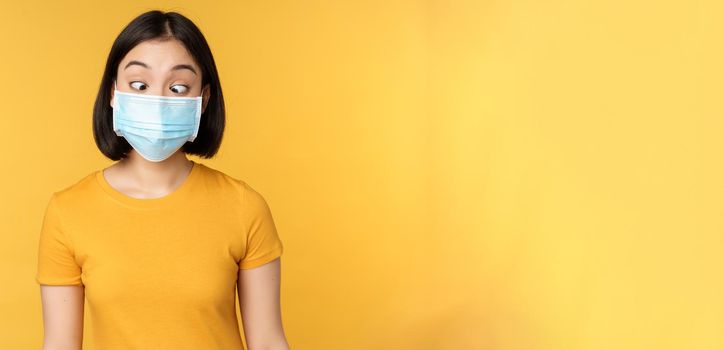 Health and people concept. Funny asian girl squinting, looking at her face medical mask, standing over yellow background.