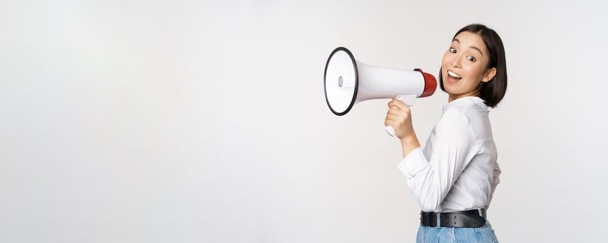 Beautiful young asian woman talking in megaphone, screams in speakerphone and smiling, making announcement, shout out information, standing over white background.