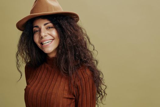 Close up portrait. Overjoyed stylish curly Latin female in brown hat, smiling at camera, laughing, saying Yeah, isolated green background. Copy space clothing fashion brands, free place for your ad.