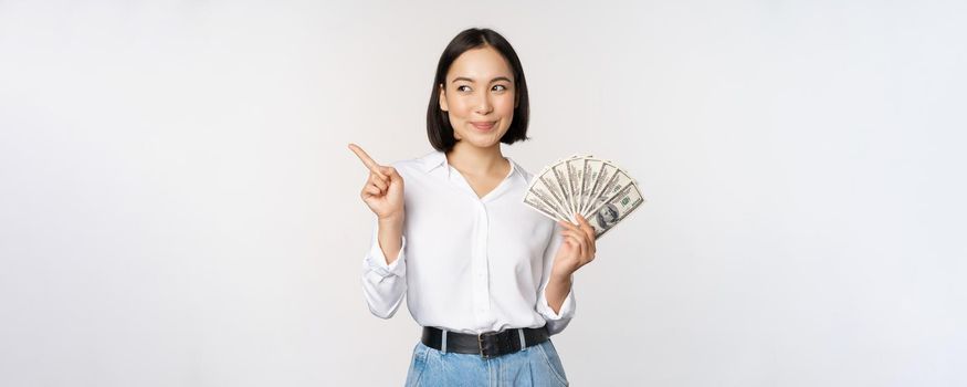 Smiling young modern asian woman, pointing at banner advertisement, holding cash money dollars, standing over white background.