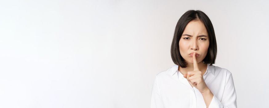 Close up portrait of young asian woman making hush, shhh shush sign, press finger to lips, dont speak, keep quiet gesture, white background.
