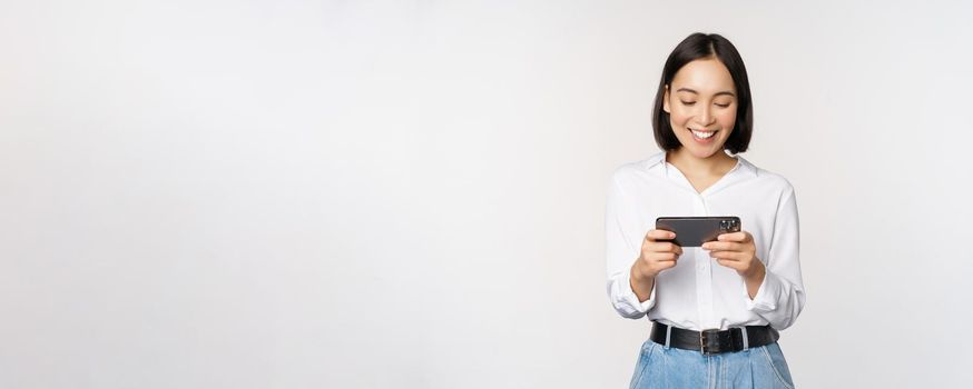 Young korean woman, asian girl playing mobile video game on smartphone, looking at horizontal phone screen, standing over white background.