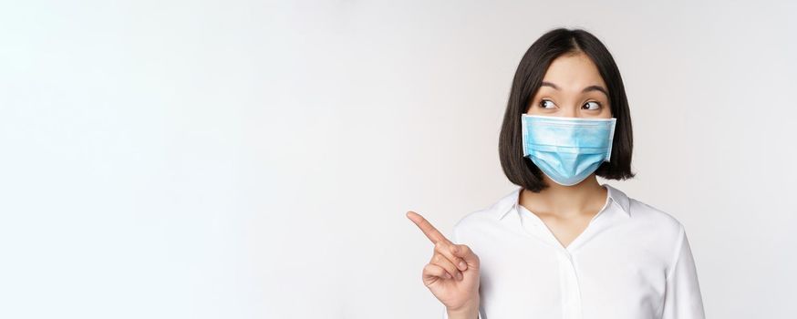 Portrait of cute asian woman in medical face mask, coronavirus protection, pointing finger left and looking intrigued at empty copy space, white background.