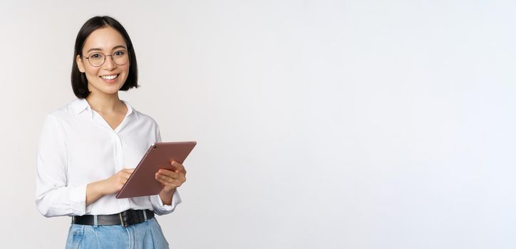 Image of young asian woman, company worker in glasses, smiling and holding digital tablet, standing over white background. Copy space