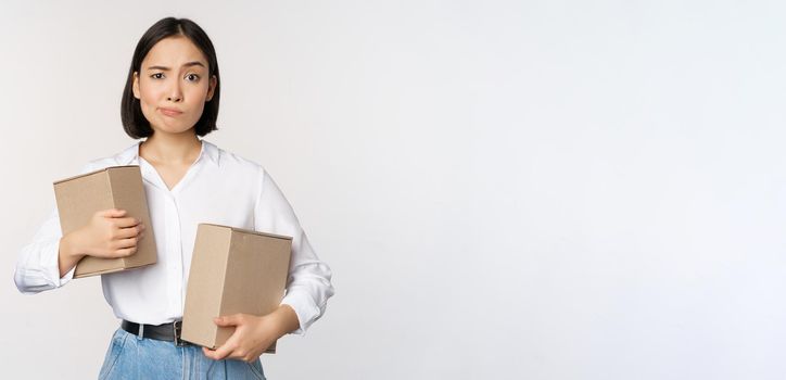 Complicated young asian woman holding two boxes, looking doubtful at camera, standing over white background puzzled.