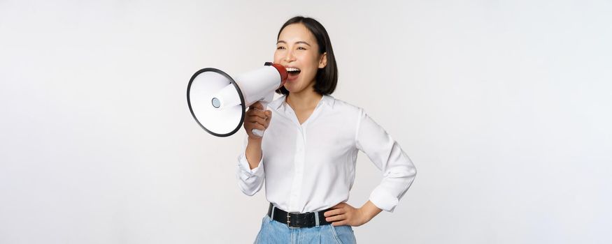 Image of young woman, korean activist, recruiter screaming in megaphone, searching, shouting at loudspeaker, standing over white background.