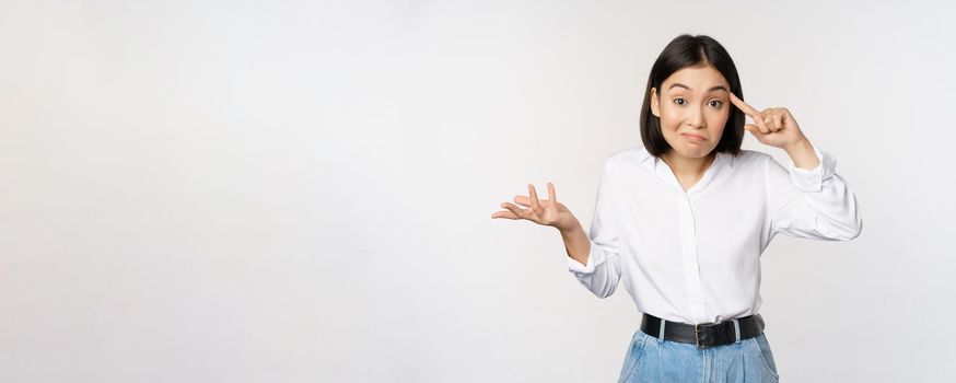 Are you insane. Portrait of frustrated young asian woman, roll finger near head and srugging, someone crazy or stupid, standing over white background.