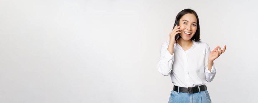 Friendly smiling asian woman talking on phone, girl on call, holding smartphone and laughing, speaking, standing over white background.