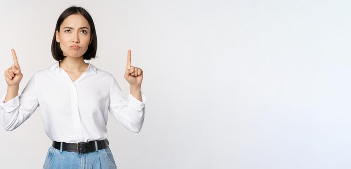 Skeptical businesswoman, asian office manager, pointing fingers up and grimacing doubtful, hesitating, standing over white background.