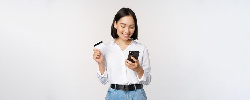 Online shopping concept. Korean woman holding credit card and looking at smartphone app, buying, order delivery in mobile phone application, standing over white background.