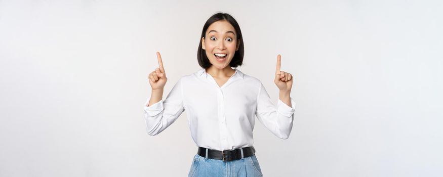 Enthusiastic business woman, asian female model pointing fingers up and smiling, making announcement, showing logo banner on top, white background.