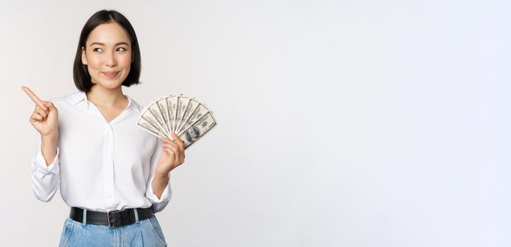 Smiling young modern asian woman, pointing at banner advertisement, holding cash money dollars, standing over white background.
