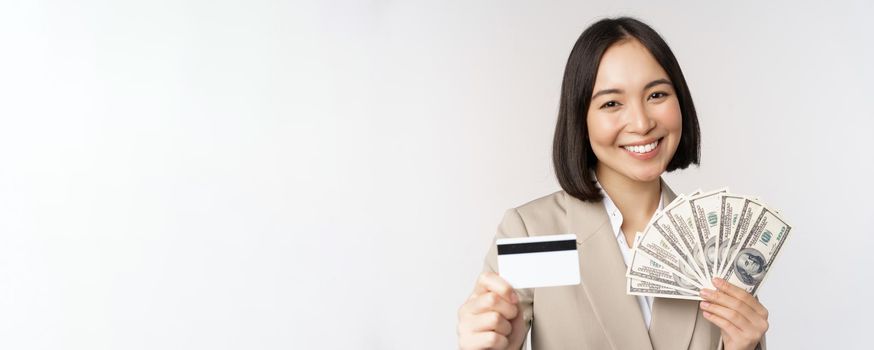 Close up of asian businesswoman, office lady showing credit card and money dollars, standing in suit over white background.