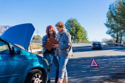 Mother and daughter standing next to the broken down vehicle checking the documents and calling the tow truck. Two friends on holiday have a car accident and have to call the mechanic in the middle of the road. Clear road on a very sunny day next to a forest with a mountain in the background. Red-haired girl calling tow truck and blonde woman looking at insurance papers.