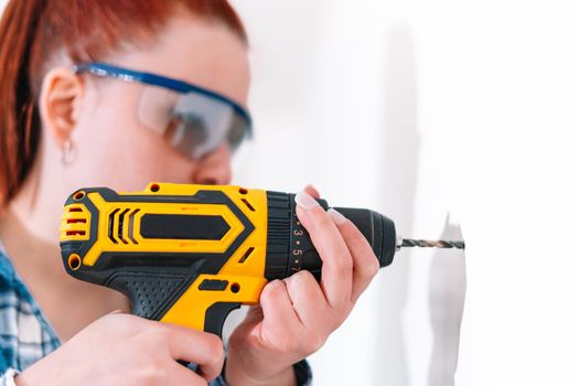 Young girl with red hair making a hole in a white wall with a drill to place a picture or a shelf. The woman is wearing blue safety goggles, a blue and white checked shirt and a yellow battery-powered drill. Her hair is tied back.