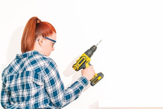 young red-haired woman with her hair tied back standing, wearing a blue chequered shirt and goggles, standing, holding a yellow drill in her right hand to hang a shelf at home on a white wall.