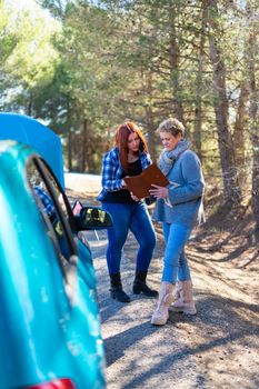 Mother and daughter looking for the documents of the car because it has broken down. Two friends checking the car insurance to call the tow truck or repair it standing on the side of a forest standing on the side of the road. Very sunny day, with a blue car. The young girl has red hair and the lady has blonde hair. Generic blue color.