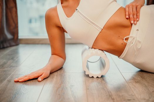 Adult athletic woman, in white bodysuit, performing fascia exercises on the floor - caucasian woman using a massage foam roller - a tool to relieve tension in the back and relieve muscle pain - the concept of physiotherapy and stretching training.