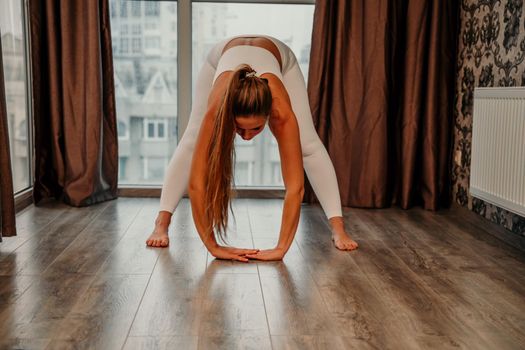 Middle aged athletic attractive woman practicing yoga. Works out at home or in a yoga studio, sportswear, white pants and a full-length top indoors. Healthy lifestyle concept.