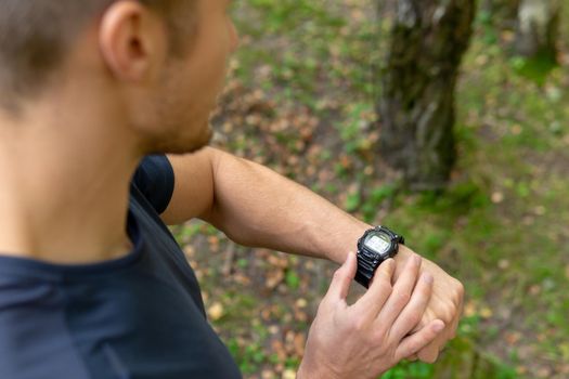 A man looks at the clock time in the park outdoors, around the forest, oaks green grass young enduring athletic athlete. healthy runner fitness trail training person outside. energy stretching stretches