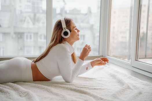 Side view portrait of relaxed woman listening to music with headphones lying on carpet at home. She is dressed in a white tracksuit