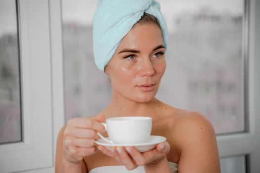 Young serene relaxed woman in spa bath towel drinking hot beverage tea coffee after taking shower bath at home. Beauty treatment, hydration concept