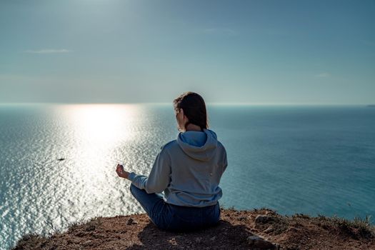 Woman tourist enjoying the sunset over the sea mountain landscape. Sits outdoors on a rock above the sea. She is wearing jeans and a blue hoodie. Healthy lifestyle, harmony and meditation.