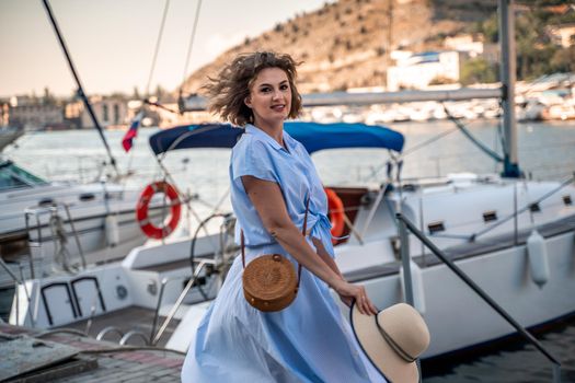 A young happy woman in a blue dress and hat stands near the seaport with luxury yachts. Travel and vacation concept.