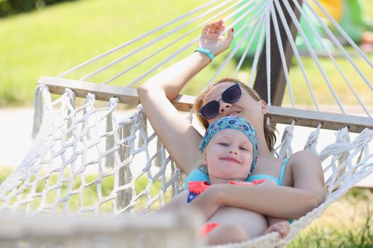 Portrait of resting mother embrace daughter, girls lay on hammock in shadow, enjoy life, time with family. Girls in swimsuits, holiday. Chilling concept