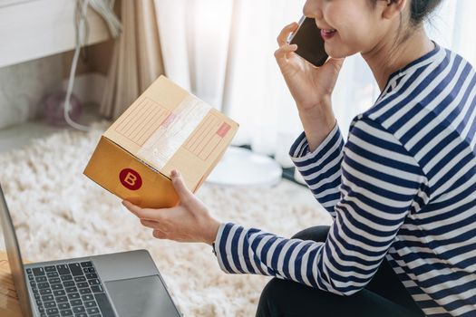 Online merchandising business idea, beautiful girl holding parcel box and talking on the phone while using computer to input Track And Trace parcel number to customers