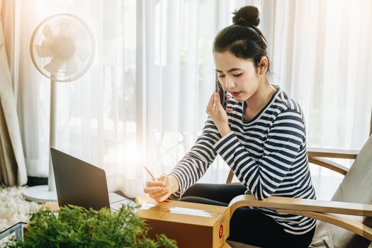 Online merchandising business idea, beautiful girl talking on the phone and holding a parcel box ready to use a computer to enter the Track And Trace parcel number to the customer