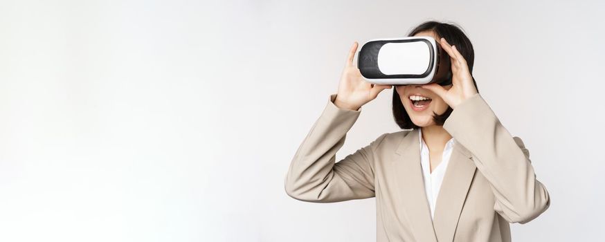 Amazed business woman in suit using virtual reality glasses, looking amazed in vr headset, standing over white background.