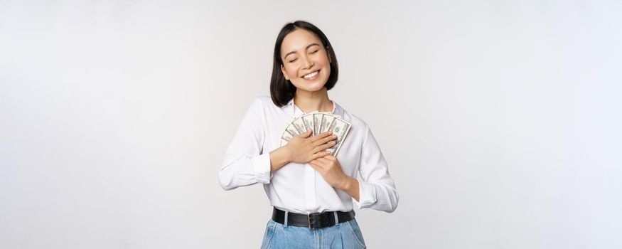 Happy asian woman hugging money dollars and smiling satisfied, standing over white background. Copy space