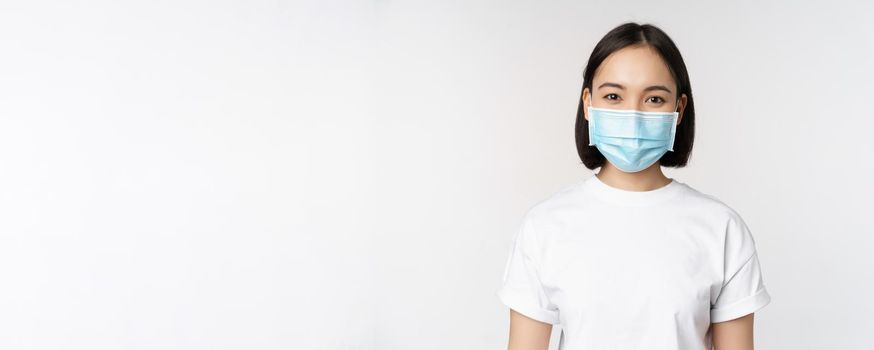 Health and covid pandemic concept. Smiling asian girl wearing medical face mask, looking happy and confident, protecting herself from coronavirus, white background.