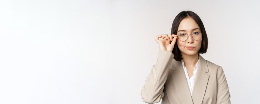 Professional asian businesswoman in glasses, looking confident at camera, standing in power pose against white background.