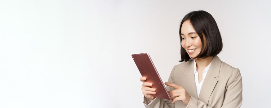 Image of asian businesswoman using digital tablet, looking at gadget and smiling, working, standing against white background.