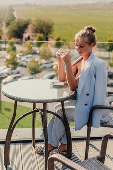A middle-aged woman sits in a street cafe overlooking the mountains at sunset. She is dressed in a blue jacket and drinks coffee admiring the nature. Travel and vacation concept