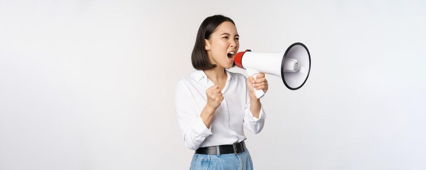 Enthusiastic asian woman, girl activist shouting at protest, using megaphone, looking confident, talking in loudspeaker, protesting, standing over white background.