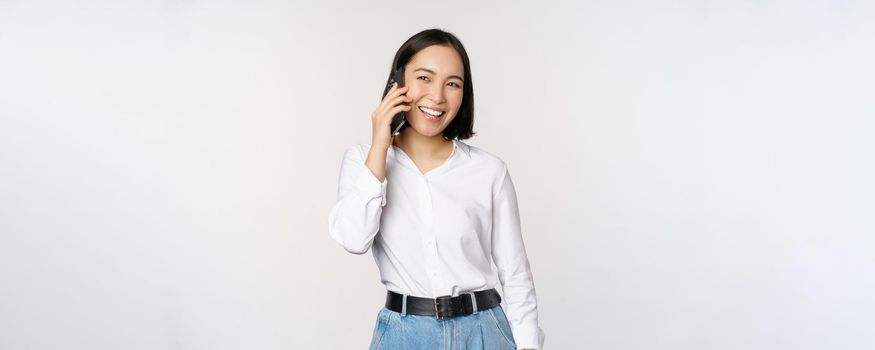 Friendly smiling asian woman talking on phone, girl on call, holding smartphone and laughing, speaking, standing over white background.