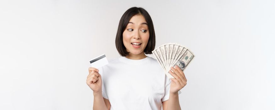 Portrait of asian woman holding money, dollars and credit card, looking impressed and amazed, standing in tshirt over white background.