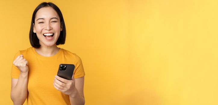 Joyful asian woman celebrating, holding mobile phone, winning, achieve goal on smartphone, standing over yellow background.