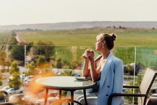 A middle-aged woman sits in a street cafe overlooking the mountains at sunset. She is dressed in a blue jacket and drinks coffee admiring the nature. Travel and vacation concept