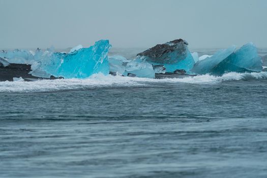 Large blue icebergs over the black sand beach beaten by the breaking waves